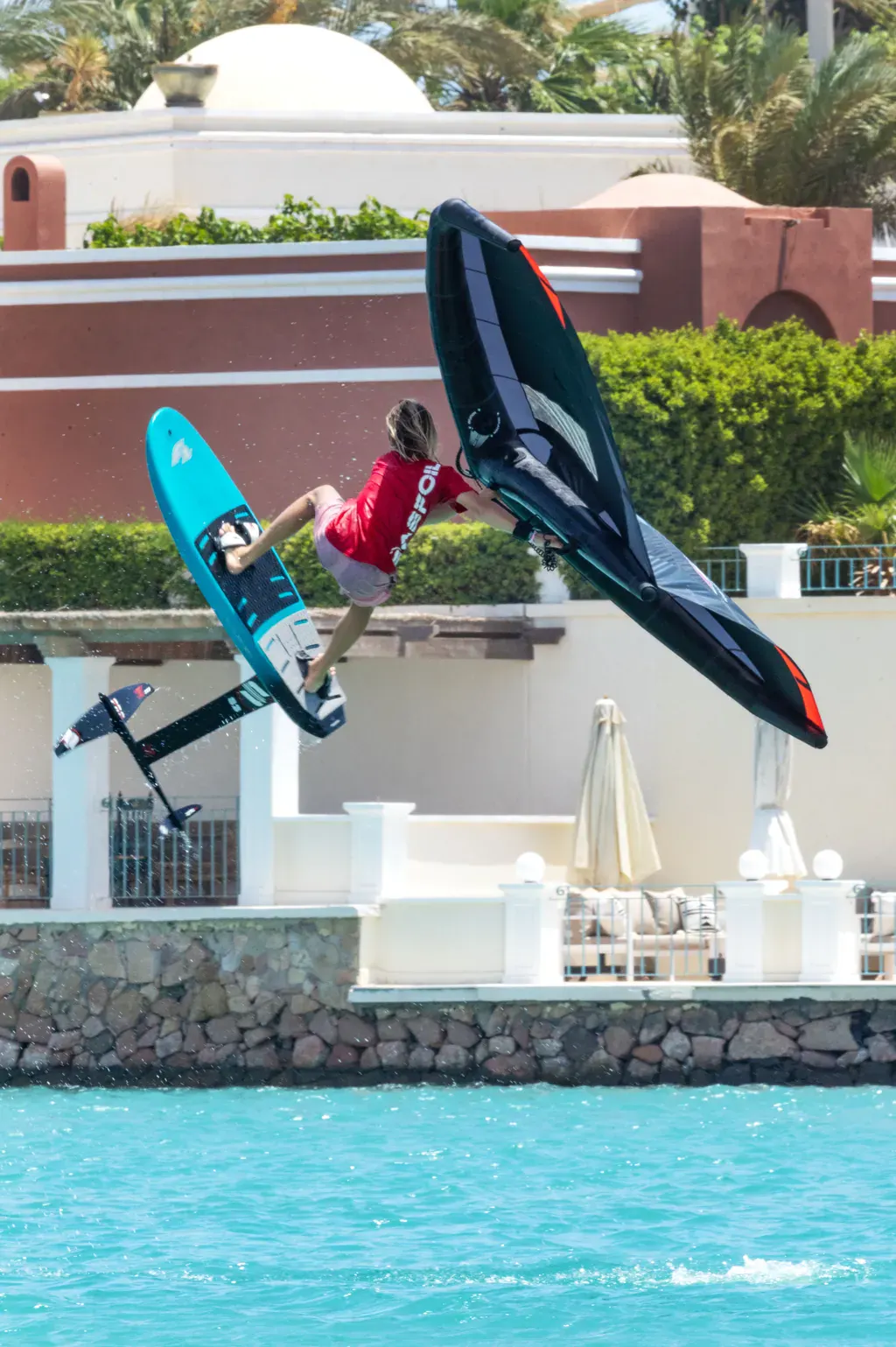 Finn Flügel airborne on a wing foil board, gripping his wing, in front of a waterfront villa with palm trees