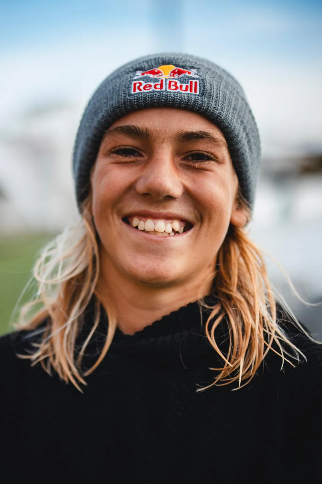 Finn Flügel smiling in a grey Red Bull beanie, his long blond hair falling over a black top