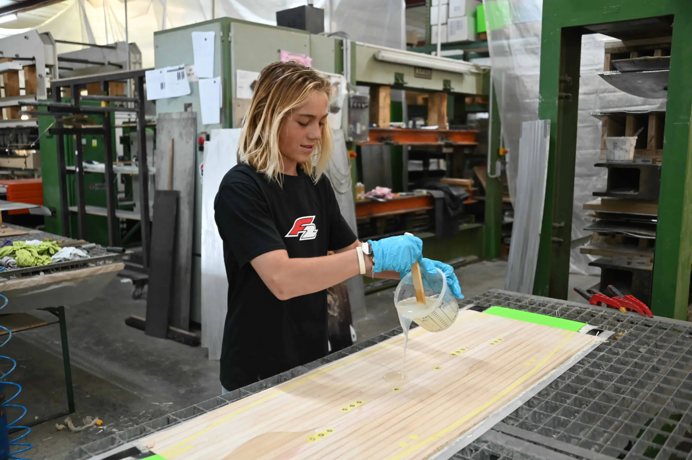 Finn Flügel, in an F2 T-shirt and blue gloves, pours resin onto a kiteboard in a board-building workshop
