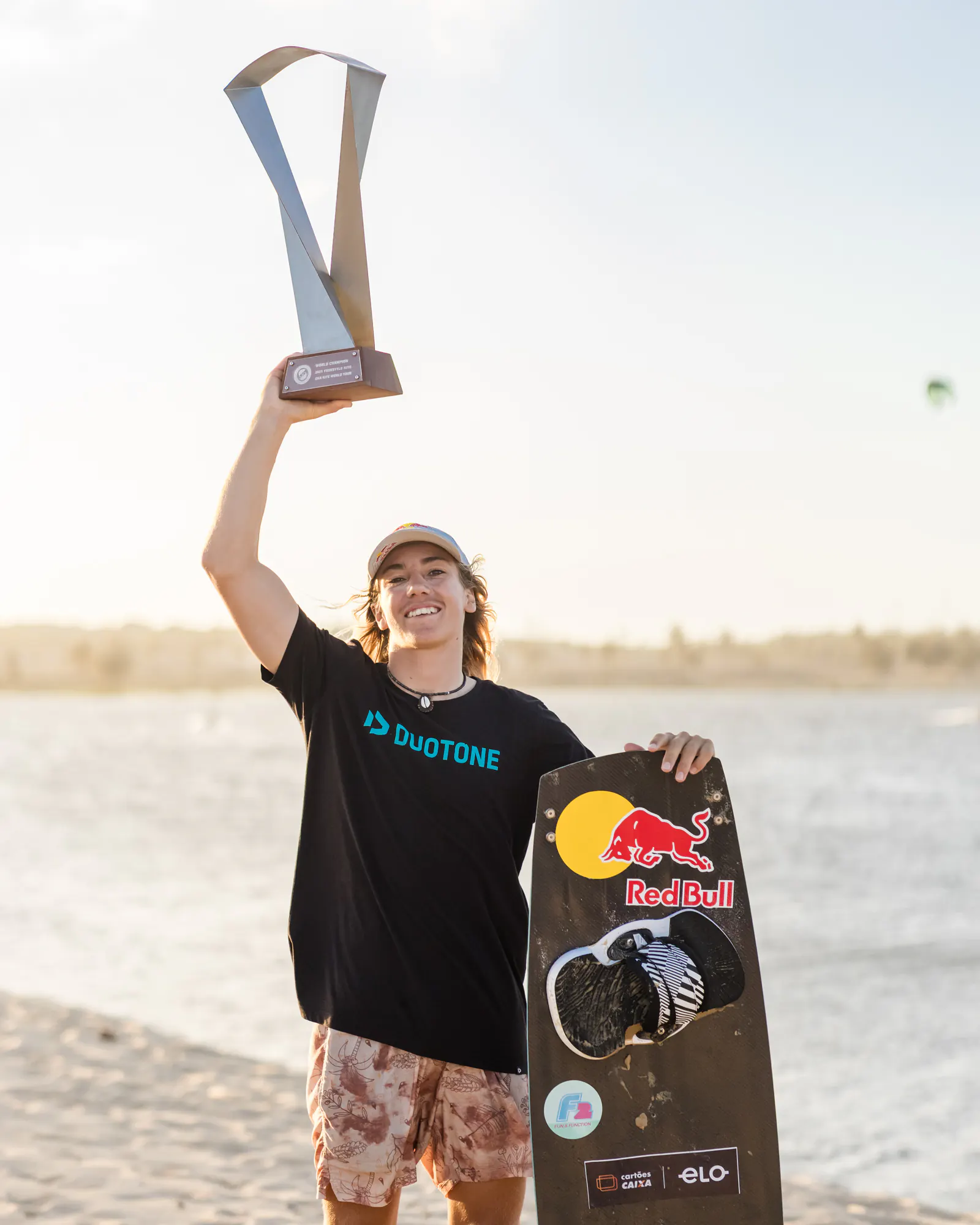 Finn Flügel raises a trophy above his head on a beach in Brazil, his Red Bull board at his side, in a Duotone T-shirt