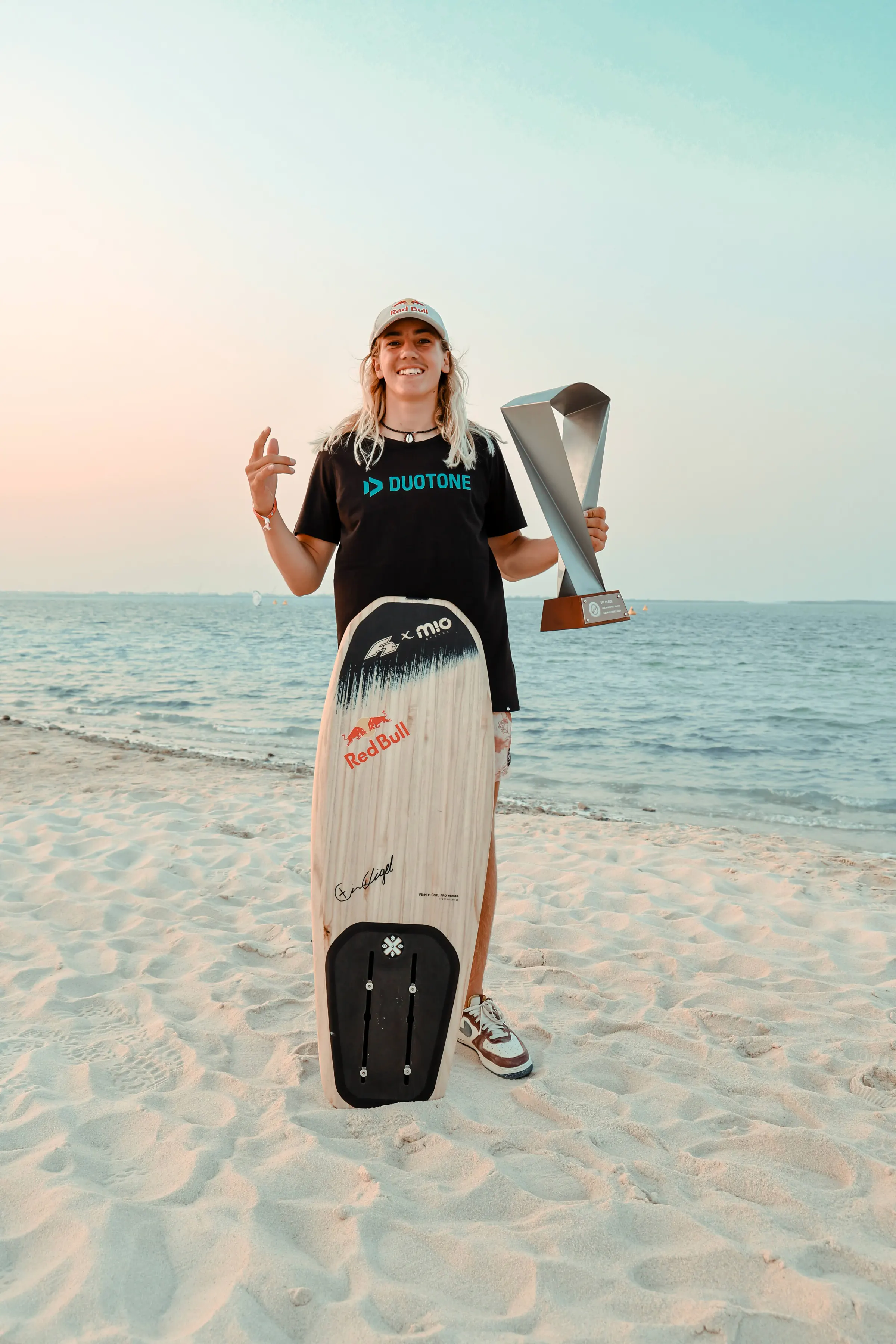 Finn Flügel on the beach at sunset with a trophy and his foil board, wearing a Red Bull cap and Duotone T-shirt
