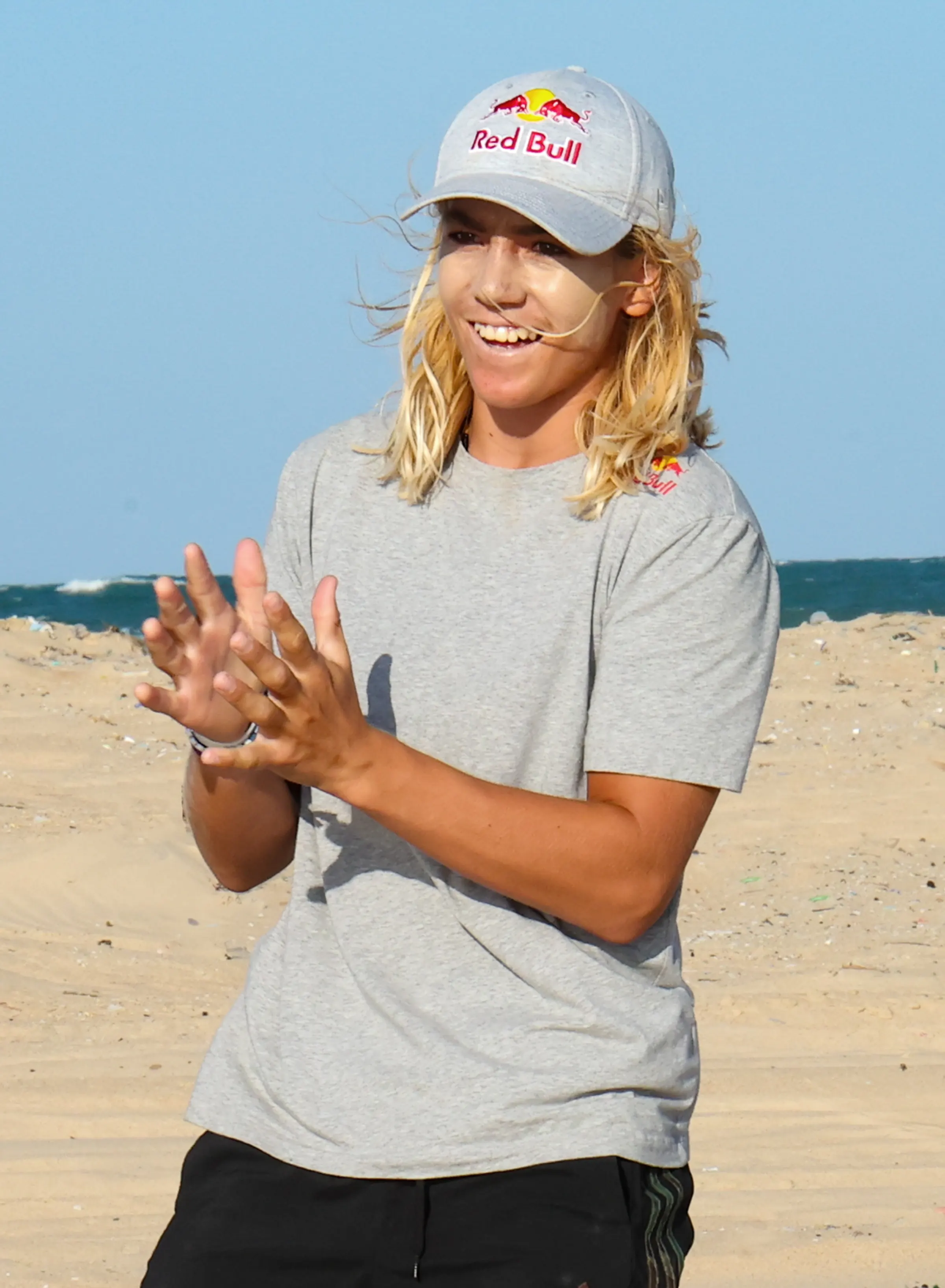 Finn Flügel smiles and claps on a sandy beach in a Red Bull cap and grey T-shirt, with the sea behind him