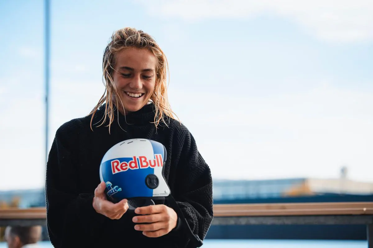 Finn Flügel, wet-haired and smiling, holds a Red Bull helmet in a black fleece top by the waterfront