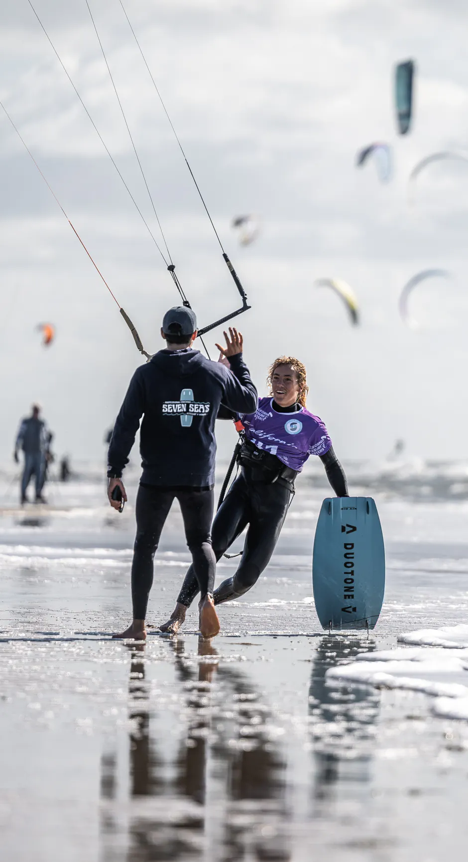 Finn in a purple competition jersey high-fives a man by the water, lots of kites in the sky behind them