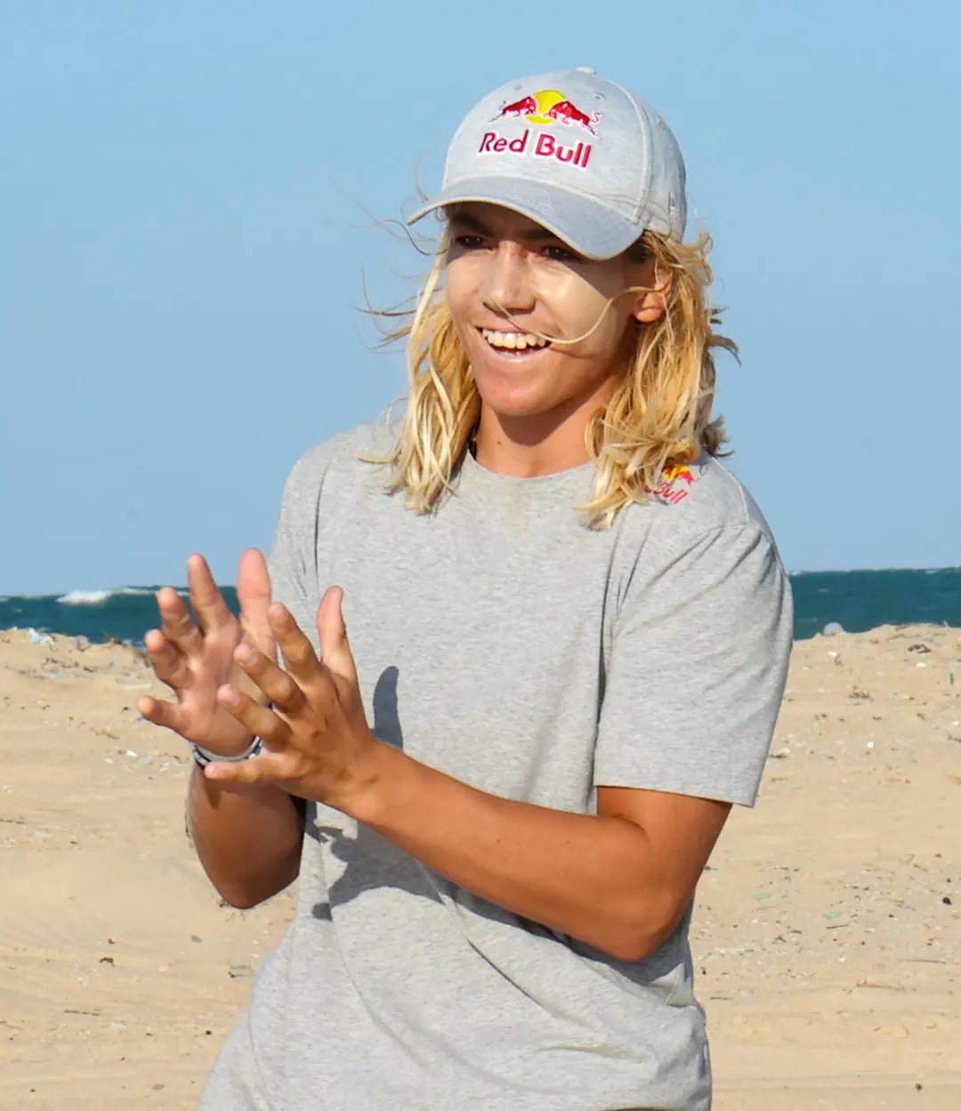 Finn in a Red Bull cap laughing and clapping on the beach, the sea behind him