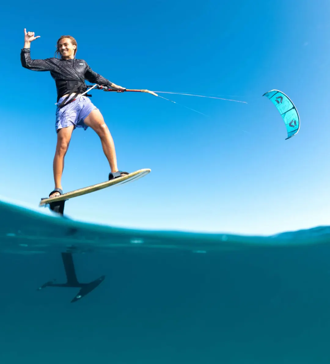 Finn on a foilboard above clear water, one hand raised in a shaka