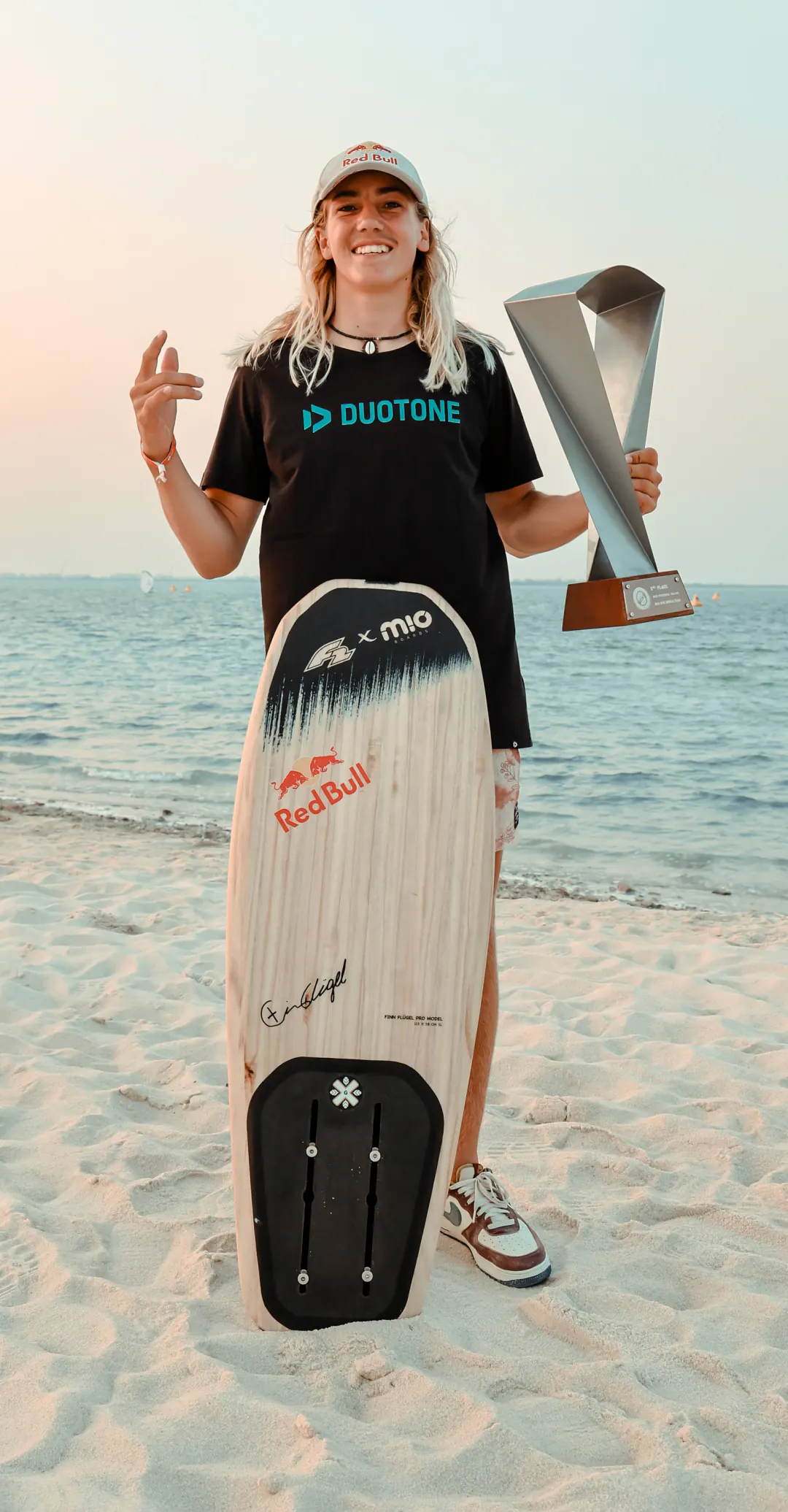 Finn on the beach at sunset with a trophy and a foilboard, in a Red Bull cap and a Duotone T-shirt