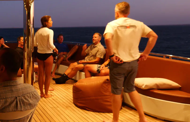 Guests sitting in the lounge on deck in the evening, the sea behind them