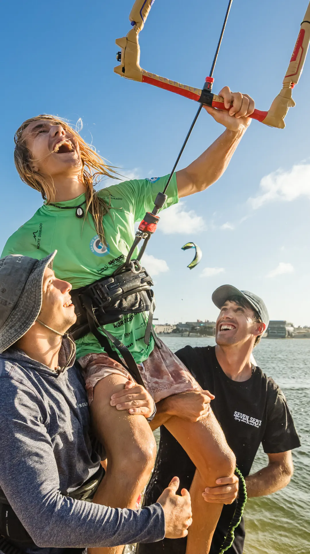 Finn, laughing, is carried on shoulders on the beach and holds his kite bar up high