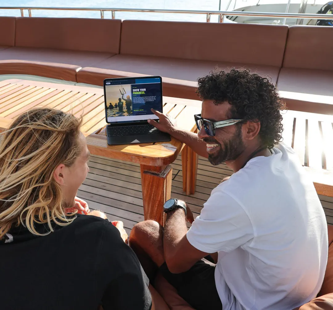 Finn and Islam sit on deck in front of a tablet; Islam, laughing, points at the screen