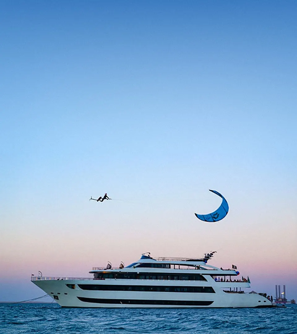 Finn in mid-air above a safari yacht at dusk, his blue kite high above the sea